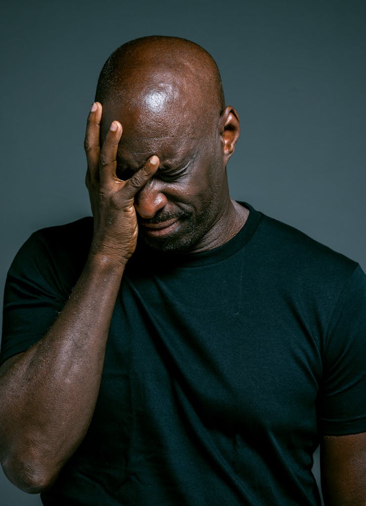 Close-up of a man in distress holding his head with eyes closed in a black shirt.