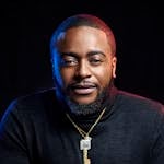 Close-up portrait of a man with a beard wearing a turtleneck and necklace against a dark background.